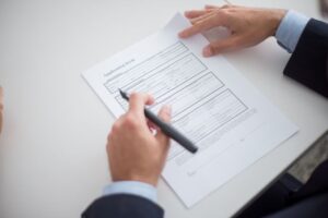 Close-up of a person in a business suit holding a pen, reviewing and preparing to sign an official application form on a white desk.