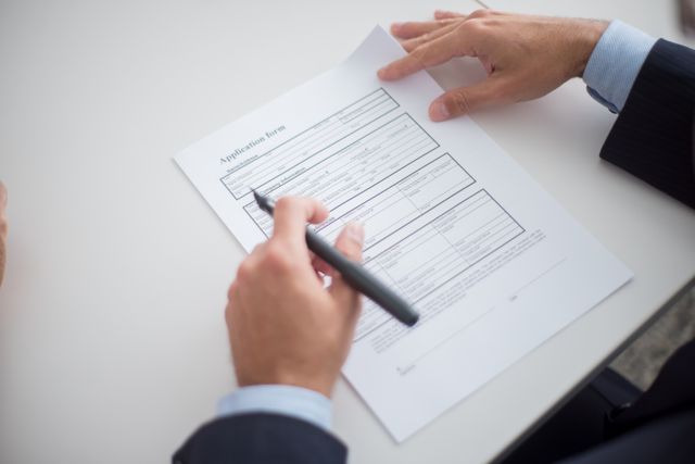 Close-up of a person in a business suit holding a pen, reviewing and preparing to sign an official application form on a white desk.