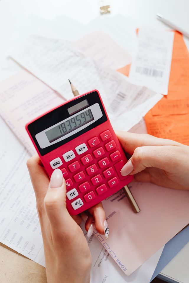 A top-down view of a person using a red calculator to tally expenses amidst a scattered pile of paper bills and receipts on a white desk.