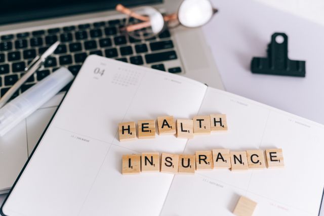 Scrabble tiles spelling "HEALTH INSURANCE" placed on an open notebook next to a laptop, eyeglasses, and a pen.