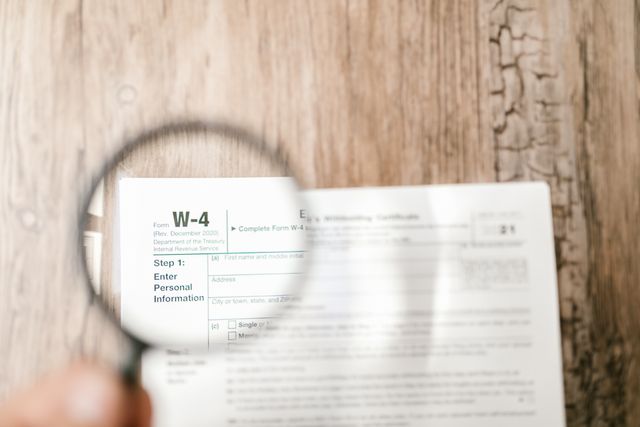 A close-up shot of a white paper W-4 tax form lying on a light-colored wooden table. A person's hand holds a round magnifying glass over the left side of the document, focusing on and enlarging the words "Form W-4" and "Step 1: Enter Personal Information." The rest of the form is slightly blurred in the background.