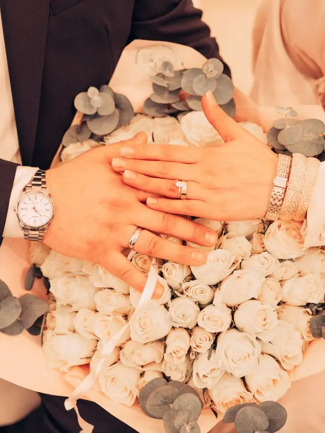 Close-up of a couple's hands with wedding rings and watches, resting on a bouquet of white roses.