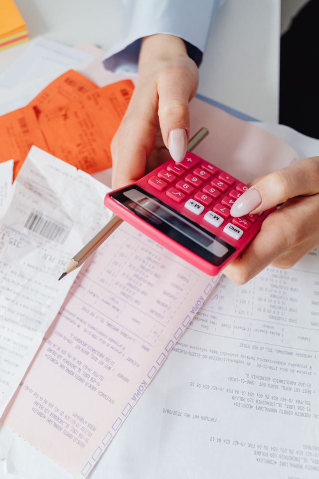 A close-up view of a person using a pink calculator to review multiple financial documents, bills, and tax invoices spread out on a desk.