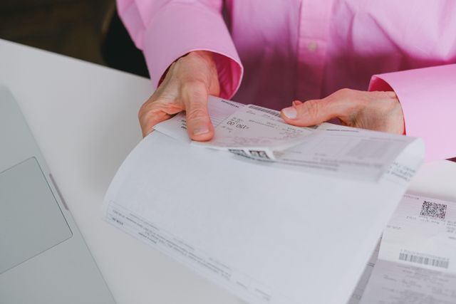 A person in a pink button-down shirt holding and reviewing folded official documents and financial statements at a white desk next to a laptop.