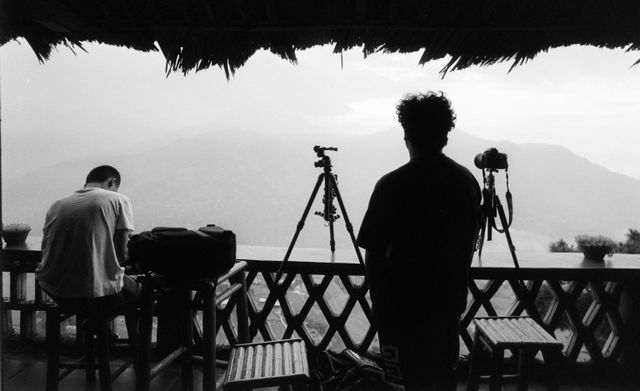 An artistic black-and-white photograph of two photographers under a thatched roof overlook, waiting to capture a misty mountain landscape. One person sits working on equipment, while another stands looking out toward the mountains next to two cameras mounted on tripods.