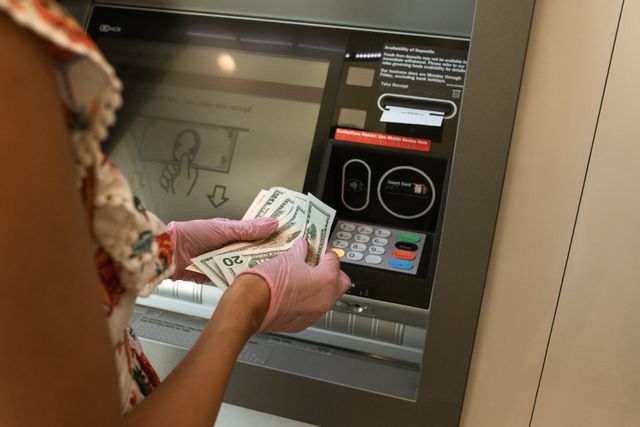A person wearing pink gloves stands in front of an ATM, counting a stack of US dollar bills, including twenty and one hundred dollar notes, preparing to make a cash deposit.