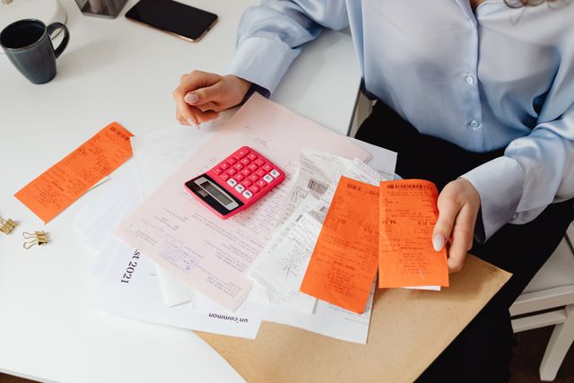 A close-up of a person sitting at a white desk reviewing a scattered pile of medical bills and orange receipts, holding a pink calculator while calculating healthcare costs.
