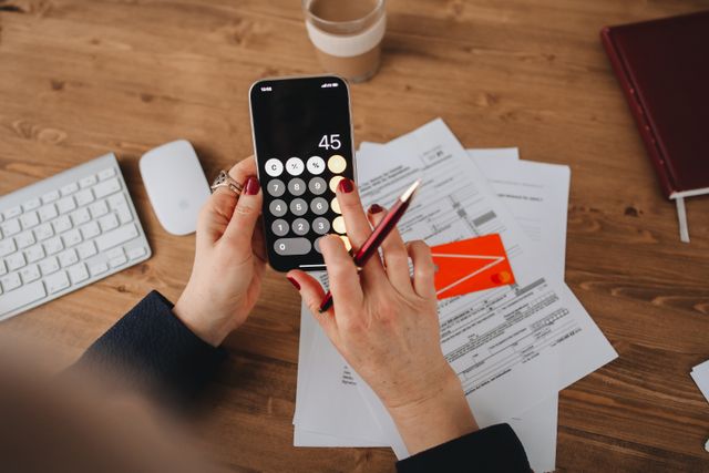 "A close-up, top-down view of a person using a calculator app on a smartphone to compute numbers, holding a pen over official financial documents and a credit card on a wooden desk."