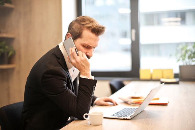 "A focused businessman in a suit sitting at his desk, looking stressed while answering a spam phone call on his smartphone next to his open laptop.