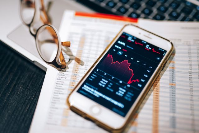 A close-up, high-angle shot of a white smartphone resting on top of a printed financial newspaper next to a pair of tortoiseshell reading glasses. The smartphone screen displays a dark-themed stock market application showing a sharp downward trend and a red line graph for Apple stock (AAPL).