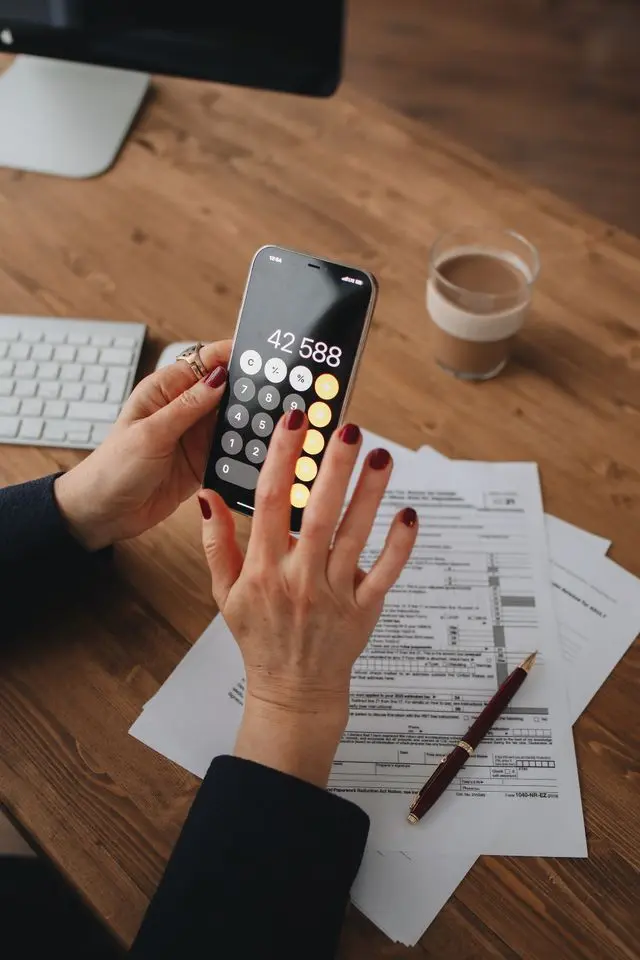 A close-up of a person holding a smartphone showing a high calculation figure of 42,588, while reviewing official tax documents and forms on a wooden desk.