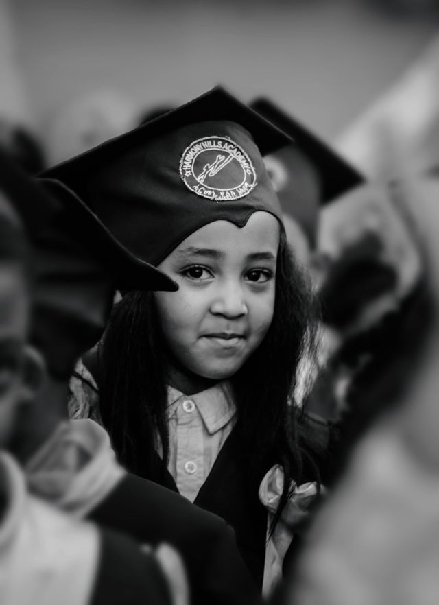 A close-up black and white photograph of a young girl wearing a graduation cap and school uniform, looking thoughtfully at the camera amidst a crowd of children.