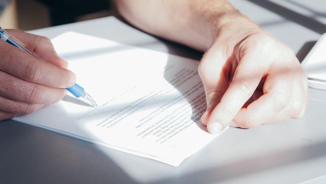 A close-up shot of a person holding a blue pen, preparing to sign or review a legal document on a sunlit desk.