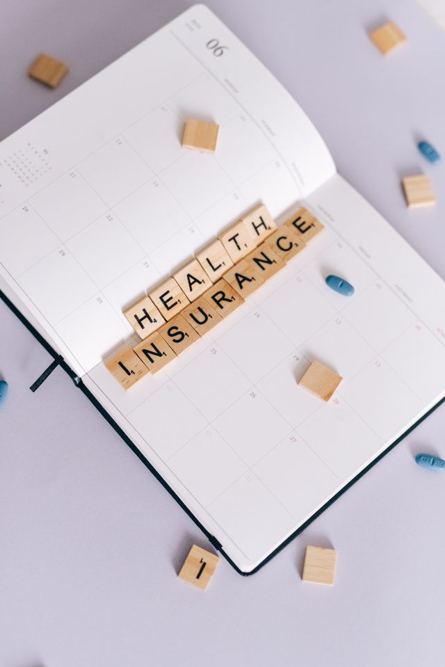 "An open monthly planner with wooden tile blocks spelling out 'HEALTH INSURANCE', surrounded by a few blue pills on a light purple surface."