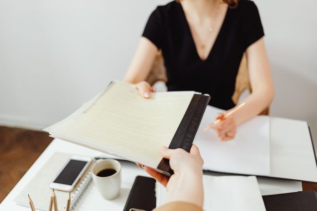 A close-up, first-person perspective of a hand handing over a legal file or document folder to a woman sitting at an office desk, illustrating the moment someone receives a lawsuit or legal notice.