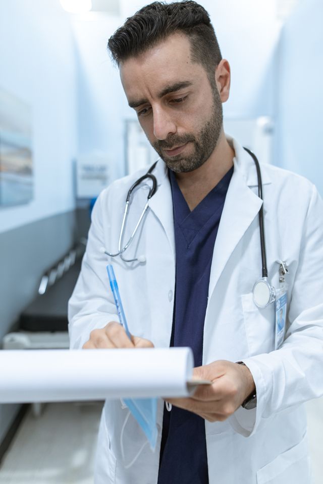 "A male doctor in a white lab coat and blue scrubs writing notes on a clipboard in a hospital hallway, representing a healthcare professional or medical billing specialist."