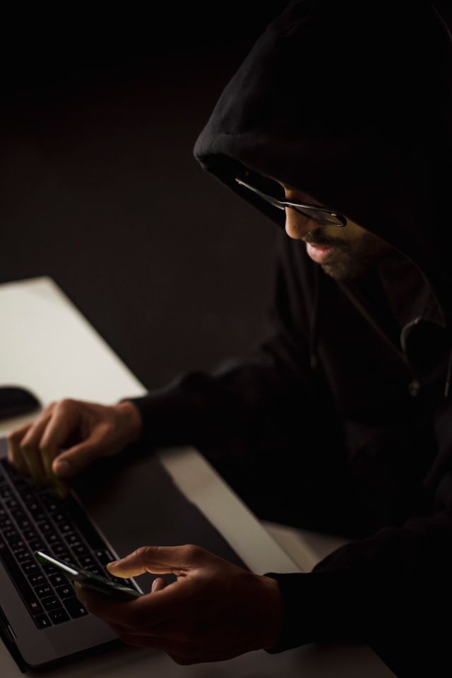 "A scammer wearing a dark hoodie using a smartphone and laptop in the dark to commit financial fraud."