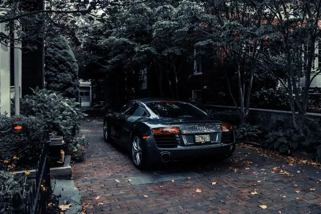 A rear-angle view of a dark grey Audi R8 sports car parked on a brick driveway, surrounded by lush green trees and foliage in a dimly lit, moody setting.