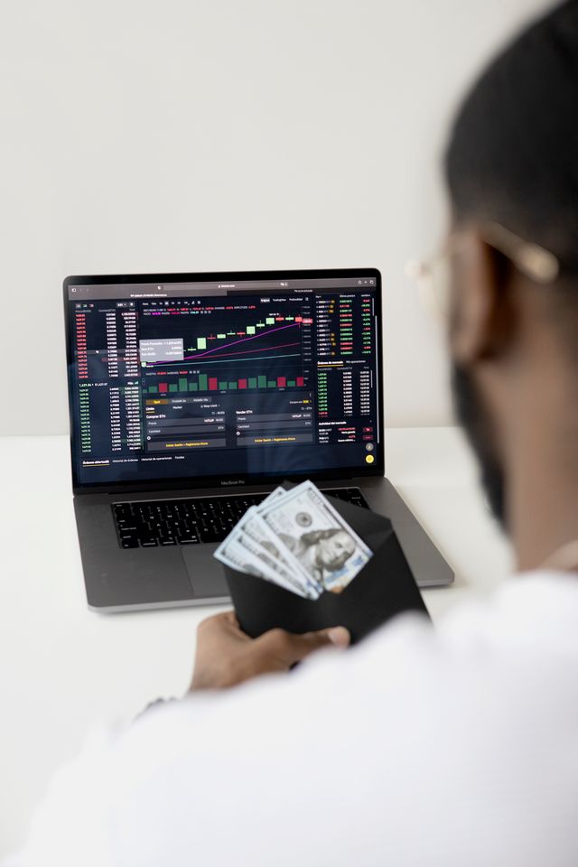 "Over-the-shoulder view of a person holding a black envelope stuffed with physical U.S. dollar bills, while looking at a laptop screen displaying complex financial trading charts and stock market data, illustrating the concept of simulated versus real trading capital."